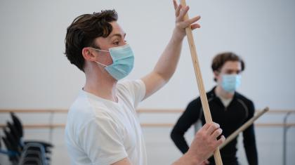 A masked choreographer in a white t-shot holds a wooden rod whilst demonstrating to the room