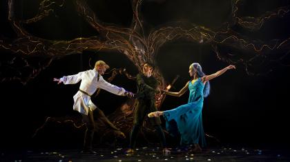 Three dancers perform in front of a large lit tree. Two dance around the other stood in the centre, holding their hands.