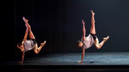 Two male dancers stand on one arm with opposite arm and foot pointed to the ceiling.