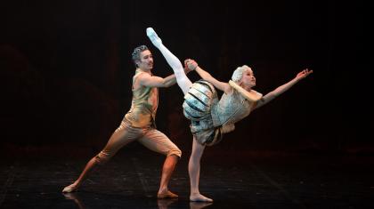 A female dancer in light, 18th century costume reaches into the distance with her leg aloft, supported from behind my a male dancer in a gold costume.