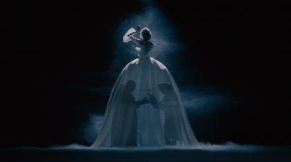 A woman in a huge white dress stands in the dark as the shadows of two men are projected onto her dress.