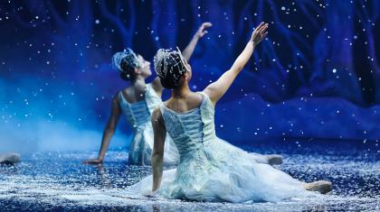 Two dancers dressed in white sit on the stage while snow falls around them