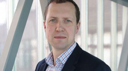 A white man with dark hair looks at the camera. He's wearing an open collared checked shirt and a dark suit jacket.