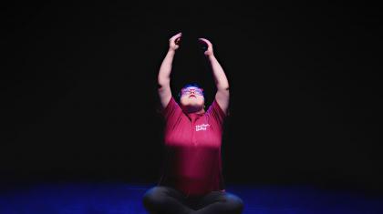 PErformer in a red T-Shirt with a darkened background looks up and reaches both of her arms straight up