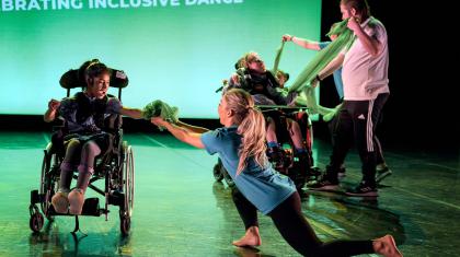 Dancer in a wheelchair reaching out and smile as someone else on their knees passes her a scrunched up length of fabric