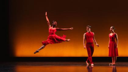 Female dancer wearing a red dress leap high with an arm stretched above her while two dancers dressed in red look on