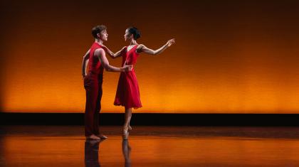 Two dancers wearing crimson, one holds the others waist and that person is en pointe, one hand on the other's shoulder