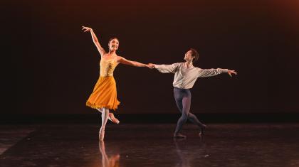 Female dancer in a orange skirt smiling as she stands on one foot en point, one leg bent at the knee behind her and an arm raised above her head while the other is held by a male dancer in white and grey