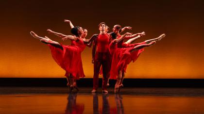 A male dancer in red is standing with arms by his side as female dancers also in red, stand arabesque around him