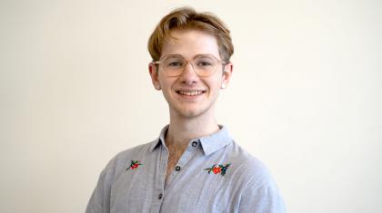 Man with fair hair, wearing glasses and a blue shirt with flower details at the shoulders smiles straight at the camera