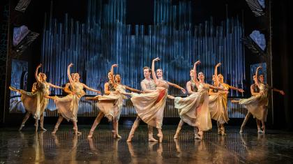 Dancers, dressed in pale gold costume, paired with the male holding the female by the waist for stability as she in en pointe ion one foot and in arabesque.