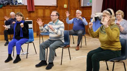 A group of people sat in plastic chairs raising their hands copying an unseen instructor