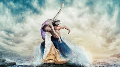 A Mermaid, pale green skin, pink-purple-white-gold outfit being held up by man on his shoulders as she leans back over them, all while being splashed by stylised crashing wavess
