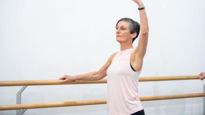 Standing at the barre with one hand holding on, a student of the Over 55s class wearing a pink top raises her other arm high while focusing in front of her