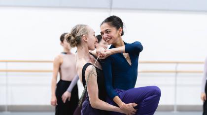 Two dancers looking at one another smiling, the one behind holding the other's leg at the thigh, the one on front reaching around playfully