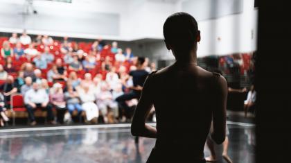 A silhouette of a dancer stood with their back to the camera while taking ballet class. On the background, a group of spectators watch from red chairs.