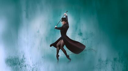 Woman holding riding crop, standing on black pointe shoes wearing a long black coat and hat