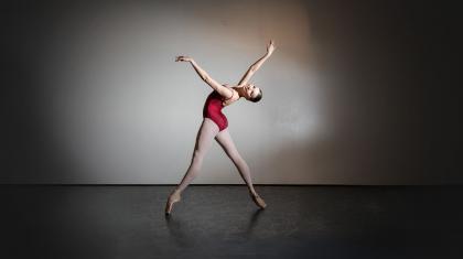 Student wearing a red leotard and white tights, en pointe, her back arched back, arms raised high, smiling at the camera