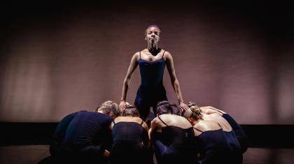 Against a dim backdrop a lone girl stands surrounded by people kneeling at her feet