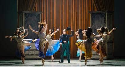 In a dressing room a group of women wearing similar dresses with different accent colours dance around Anne Lister who lifts her hat in respect to them