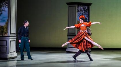 A woman in a black frock coat looks on lustfully as a woman in an orange dress is carried away while looking back at the first woman
