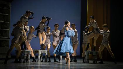 Woman in a blue dress smiles, smiling holding her hands to her chest, behind her dancers representing Anne Lister's beguiling words