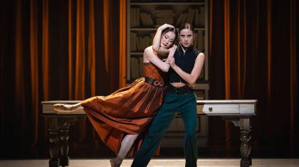 Mariana and Anne Lister stand, holding each other. Mariana in an orange dress, her eye closed as she rests her head on Anne's shoulder, Anne look straight at you