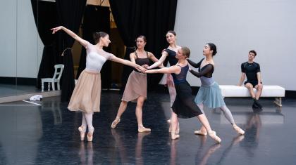 Five dancers in the rehearsal room in a circle all touching hands in the centre, one en pointe