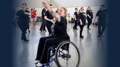 Woman in wheelchair smiling and raising her arm expressively. Young people in the background moving in unison