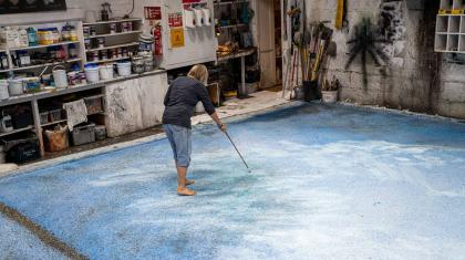 In a workshop, a person wearing a black top and jeans stands on a large canvas painting with a long paintbrush.