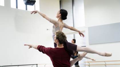 Dancer in a deep red top lifts another on his shoulder, the lifted person gracefully arches her back up, extends a hand in front of her and one leg behind
