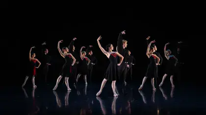 Six pair of dancers, each wearing black, the women partners with red accents on their dresses, stand posing with one hand on their hips, the other arm raise above their head