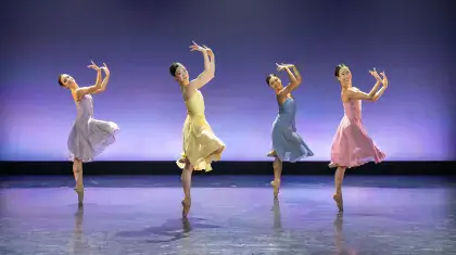 Four female dancers, one each in lilac, yellow, blue and pink, all on one foot en pointe with their arms in front of their face
