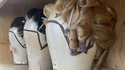 A row of 3 wigs are on display after being styled. There is one blonde and two brunette. They have rollers, pins and clips in them.