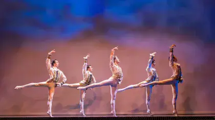 Five male dancers in white and bodies in sync, standing straight up on one foot, one hand on hip, the other arm high in the air